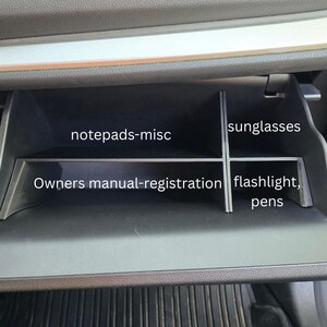 K&ouml;nnte beinhalten: Ein offenes Handschuhfach eines Autos, unterteilt in vier Bereiche. Die Bereiche sind beschriftet mit: "notepads-misc", "Owners manual-registration", "sunglasses" und "flashlight, pens". Das Fach ist schwarz und befindet sich unter einem grauen Armaturenbrett.