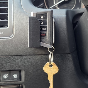 May include: A black key fob holder attached to a car's dashboard, holding a key fob with lock and hold buttons. A gold-colored key hangs from a key ring attached to the fob.