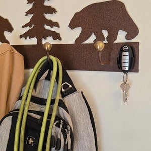 May include: A metal wall-mounted coat rack with a brown bear and pine tree design. The rack has three hooks and is decorated with gold-colored accents. A green jacket, a tan jacket, a backpack, and a green hose are hanging on the hooks. A car key with a key chain is hanging on the rightmost hook.