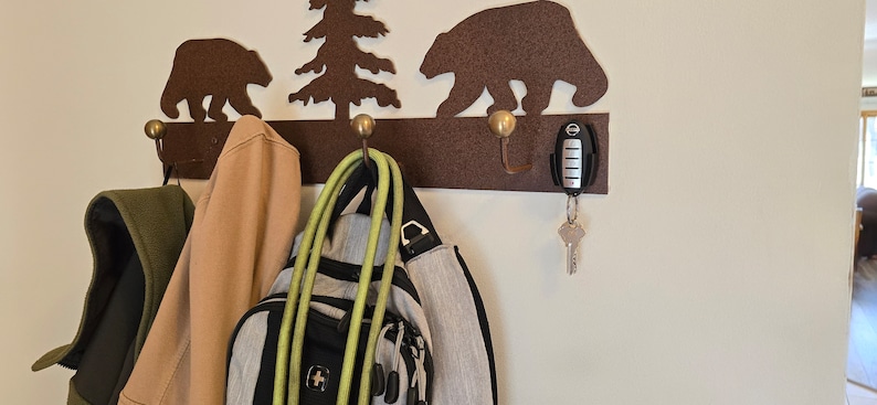 May include: A metal wall-mounted coat rack with a brown bear and pine tree design. The rack has three hooks and is decorated with gold-colored accents. A green jacket, a tan jacket, a backpack, and a green hose are hanging on the hooks. A car key with a key chain is hanging on the rightmost hook.