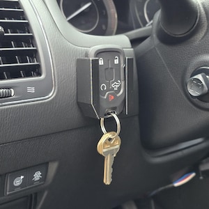 May include: Black key fob holder mounted on a car's dashboard, holding a car key. The holder is positioned near the air vent and steering wheel. The key fob has buttons for lock, unlock, and other car functions.