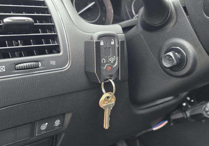 May include: Black key fob holder mounted on a car's dashboard, holding a car key. The holder is positioned near the air vent and steering wheel. The key fob has buttons for lock, unlock, and other car functions.