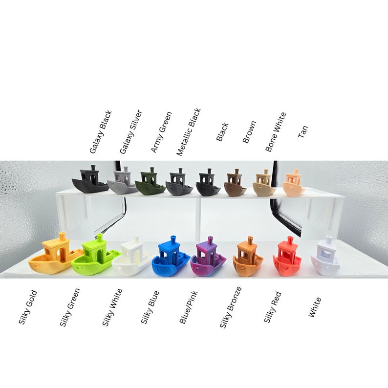 May include: A collection of small, 3D-printed toy boats in various colors, including black, brown, and white. The boats are arranged on a clear display surface, with color names like "Silky Gold" and "Silky Green" labeled below each boat. The boats are approximately 2 inches long.