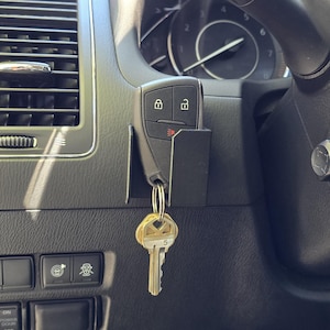 Puede incluir: Primer plano del salpicadero de un coche, con un llavero y llaves colgadas de un soporte. El salpicadero es negro con detalles plateados, incluyendo las rejillas de ventilación y los botones de control. El volante y el velocímetro son visibles en el fondo.