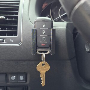 May include: A black car key fob with a key holder attached, hanging from a key ring. The key fob has buttons for locking, unlocking, and other functions. A gold-colored key is attached to the key ring.