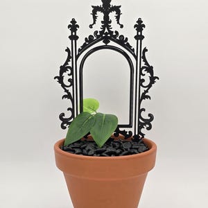 May include: A terracotta plant pot filled with black stones and green leaves, topped with a black decorative arch. The arch features an ornate design and a central opening. The plant pot is set against a white background.