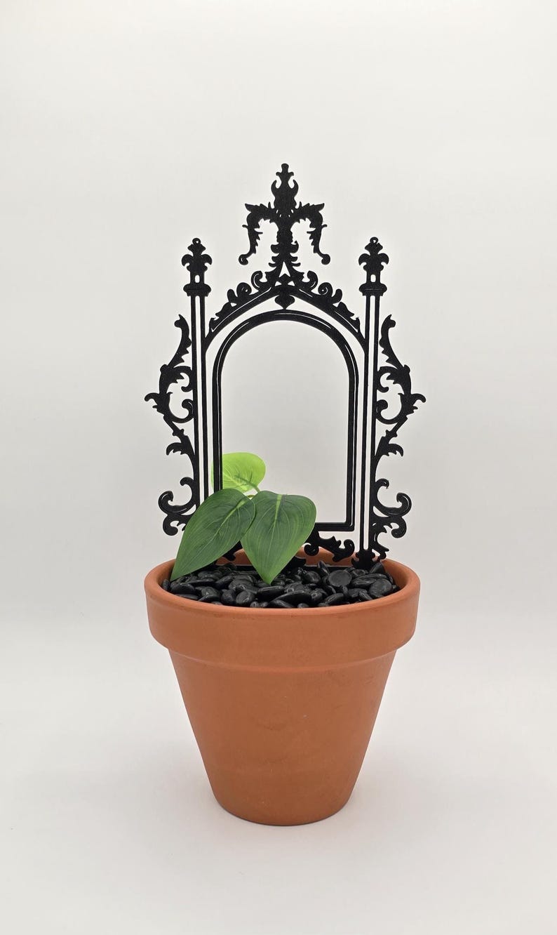 May include: A terracotta plant pot filled with black stones and green leaves, topped with a black decorative arch. The arch features an ornate design and a central opening. The plant pot is set against a white background.