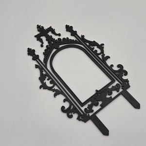 May include: Black decorative frame with ornate scrollwork and a central arched opening. The frame has two stakes at the bottom for insertion into a cake or other surface. The design is symmetrical and elegant.