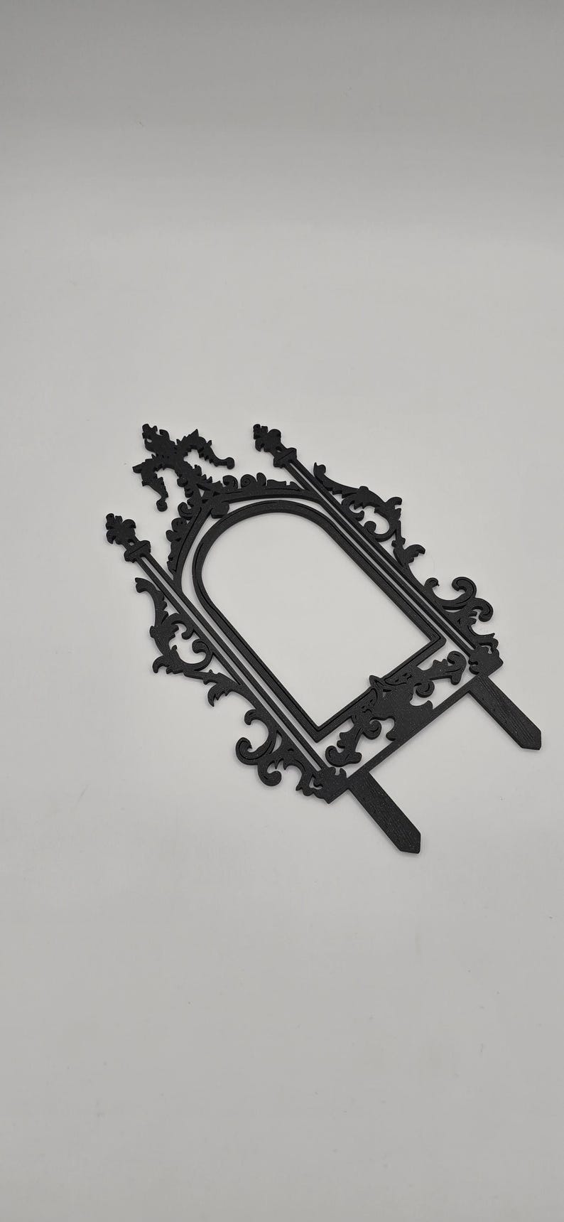 May include: Black decorative frame with ornate scrollwork and a central arched opening. The frame has two stakes at the bottom for insertion into a cake or other surface. The design is symmetrical and elegant.
