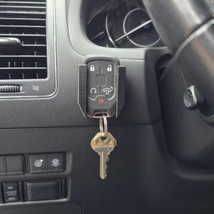 Peut inclure: Un porte-clés de voiture noir avec un porte-clés attaché, suspendu à un support noir à l'intérieur d'une voiture. Le porte-clés comporte des boutons pour verrouiller, déverrouiller et d'autres fonctions. Le tableau de bord et la grille d'aération de la voiture sont visibles.