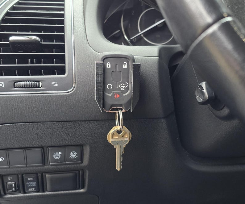 May include: A black car key fob with a key ring attached, hanging from a black holder in a car's interior. The key fob has buttons for locking, unlocking, and other functions. The car's dashboard and air vent are visible in the background.