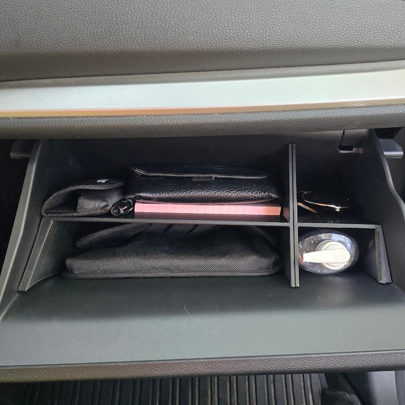 K&ouml;nnte beinhalten: Ein offenes Handschuhfach im Auto mit verschiedenen Gegenst&auml;nden. Eine schwarze Ledergeldb&ouml;rse, ein rosa Notizbuch, eine Sonnenbrille und eine Handdesinfektionsmittelflasche sind sichtbar. Das Fach ist in Abschnitte unterteilt.