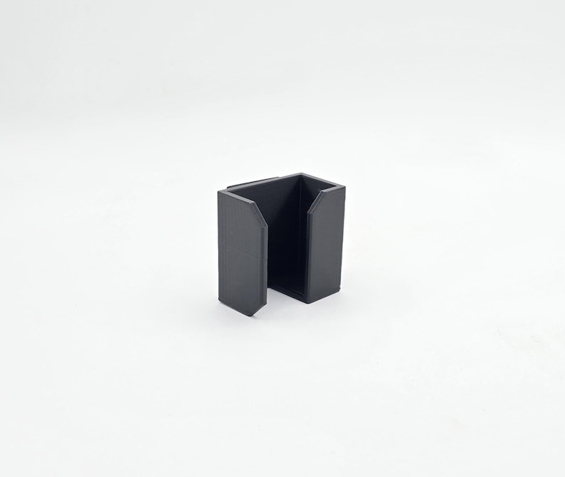 May include: A black, open-sided, rectangular container with a squared-off design. The container has a matte finish and is set against a white background. The object appears to be a holder or organizer.
