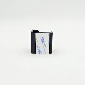 May include: A small, black, square-shaped object with a cutout on one side. A white label with blue text and the "3M" logo is affixed to the front. The label reads "Double Coated Tape Tissue".