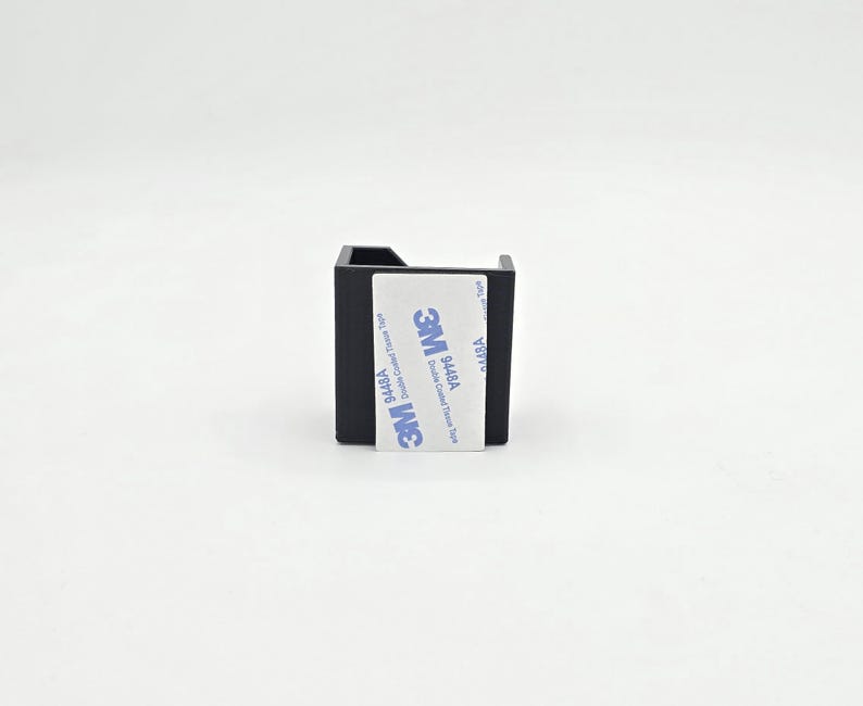 May include: A small, black, square-shaped object with a cutout on one side. A white label with blue text and the "3M" logo is affixed to the front. The label reads "Double Coated Tape Tissue".