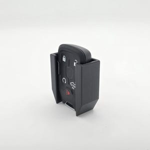 May include: A black car key fob is held in a black plastic holder. The key fob has several buttons and symbols, including a lock, unlock, and trunk release. The holder is designed to protect the key fob.
