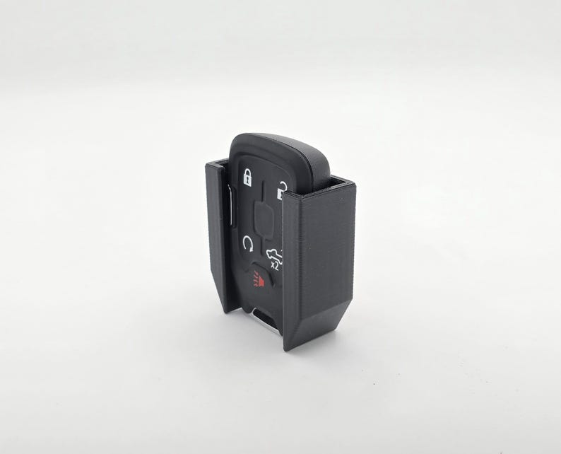 May include: A black car key fob is held in a black plastic holder. The key fob has several buttons and symbols, including a lock, unlock, and trunk release. The holder is designed to protect the key fob.