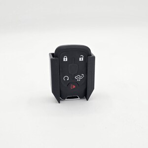 May include: A black car key fob with a protective black holder. The key fob has buttons for lock, unlock, and other vehicle functions. The holder has a rectangular shape with open sides.