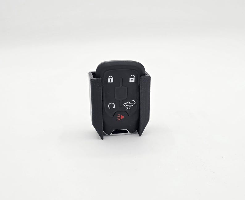 May include: A black car key fob with a protective black holder. The key fob has buttons for lock, unlock, and other vehicle functions. The holder has a rectangular shape with open sides.