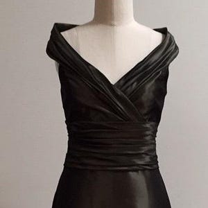 May include: A black, knee-length dress displayed on a mannequin. The dress features an off-the-shoulder neckline with draped fabric and a cinched waist. The skirt has a slight A-line shape, and the fabric has a subtle sheen.