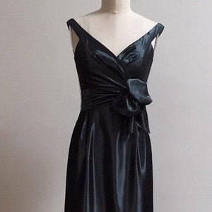 May include: A black satin evening gown displayed on a mannequin. The dress features a V-neckline, shoulder straps, and a bow detail at the waist. The floor-length skirt has a slight sheen.