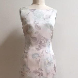 May include: A sleeveless, knee-length dress with a floral pattern. The dress features a light pink background with a repeating pattern of large, pale green and gray roses and leaves. The dress is displayed on a mannequin.