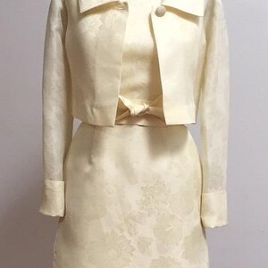 May include: A yellow dress and matching jacket set with a floral pattern. The dress has a fitted bodice and a skirt that falls to the knee. The jacket is cropped and has a collar.