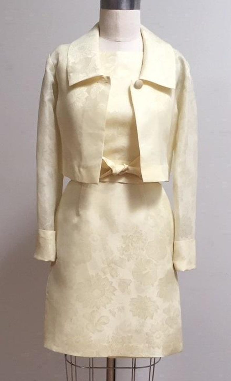 May include: A yellow dress and matching jacket set with a floral pattern. The dress has a fitted bodice and a skirt that falls to the knee. The jacket is cropped and has a collar.