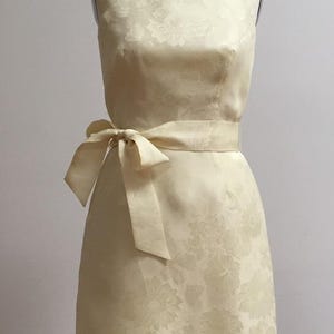 May include: A sleeveless, pale yellow dress with a floral pattern. The dress has a fitted bodice and a slightly flared skirt. A matching sash is tied at the waist.