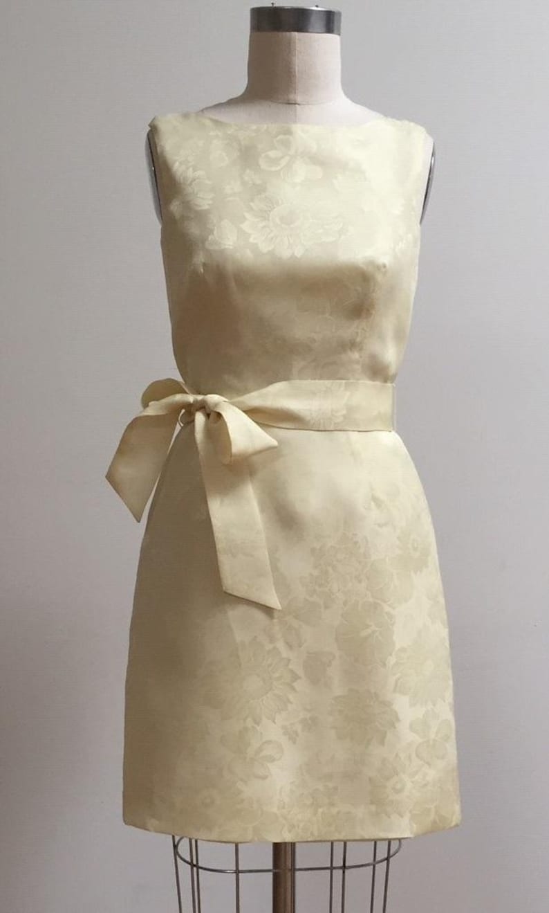 May include: A sleeveless, pale yellow dress with a floral pattern. The dress has a fitted bodice and a slightly flared skirt. A matching sash is tied at the waist.