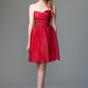 May include: A strapless, knee-length red dress. The bodice is ruched and fitted, with a defined waist and a flared skirt. The dress is paired with silver heels. The background is a solid gray.