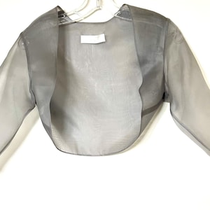 Puede incluir: Bolero gris transparente con mangas largas, colgado de una percha blanca. La tela ligera es semitransparente, revelando la forma de la prenda. El bolero tiene un frente redondeado y una pequeña etiqueta cerca del cuello.