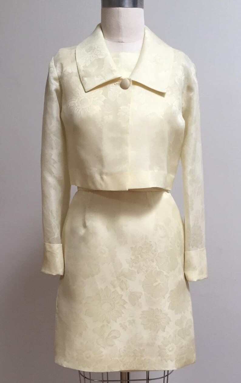 May include: A vintage ivory-colored dress suit with a cropped jacket and a skirt. The fabric has a floral pattern.