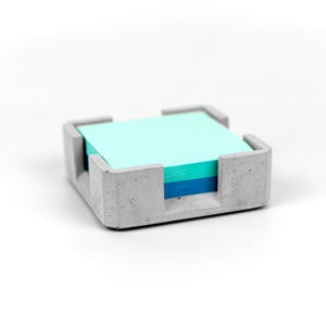 Könnte beinhalten: Ein quadratischer, grauer Schreibtisch-Organizer aus Beton, der einen Stapel bunter Haftnotizen enthält. Die Notizen sind in Blau- und Grüntönen gehalten. Der Organizer hat ein minimalistisches Design mit quadratischer Basis und vier Eckstützen.