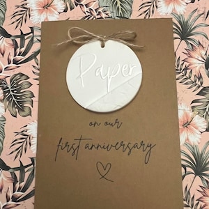 May include: A brown card with the words "Paper on our first anniversary" and a heart symbol. A white round ornament with the word "Paper" is attached to the card with twine.