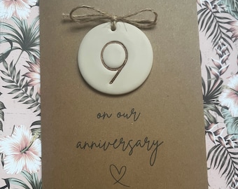 Könnte beinhalten: Eine braune Karte mit dem Text "on our anniversary" und einem Herzsymbol. Ein kleines, rundes, beigefarbenes Keramikornament mit der Zahl "9" darauf ist mit einer Schnur an der Karte befestigt.