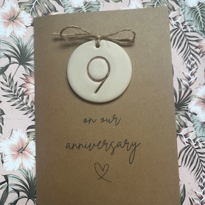 May include: A brown card with the text "on our anniversary" and a heart symbol. A small, round, beige ceramic ornament with the number "9" inscribed on it is attached to the card with twine.