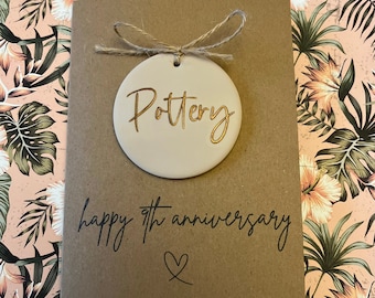 Könnte beinhalten: Eine braune Karte mit einem cremefarbenen Kreis, der mit einer Schnur befestigt ist. Der Kreis trägt die Aufschrift "Pottery" in goldenen Buchstaben. Die Karte lautet "happy 9th anniversary" mit einem Herzsymbol.
