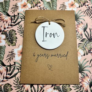 May include: A brown card with a white circle attached to it with twine. The circle has the word "Iron" written on it in black. The card says "6 years married" with a heart symbol below the text.