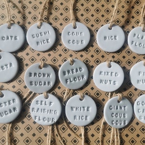 May include: A set of 16 grey ceramic labels with twine attached. Each label has a different food item written on it, such as oats, sushi rice, couscous, pasta, brown sugar, bread flour, mixed nuts, fine nuts, caster sugar, self raising flour, white rice, giant couscous, and golden caster sugar.