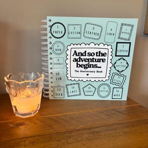May include: A light blue spiral-bound anniversary book titled And so the adventure begins... The Anniversary Book, featuring a list of sixteen anniversary themes from paper to wax, positioned next to a clear glass votive candle holder with a lit candle.