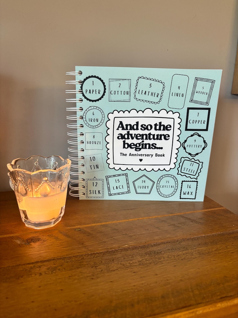 May include: A light blue spiral-bound anniversary book titled And so the adventure begins... The Anniversary Book, featuring a list of sixteen anniversary themes from paper to wax, positioned next to a clear glass votive candle holder with a lit candle.