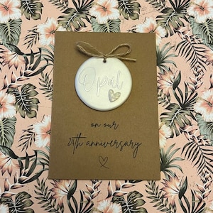 May include: A brown card with the word "Opal" written on a white circle attached to the card with twine. The card also says "on our 24th anniversary" with a small heart symbol.