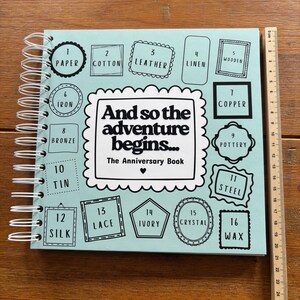 May include: A light blue spiral-bound anniversary book with the text "And so the adventure begins... The Anniversary Book" and sixteen numbered frames listing materials from 1 to 16. A wooden ruler shows the book is 28 cm in height.