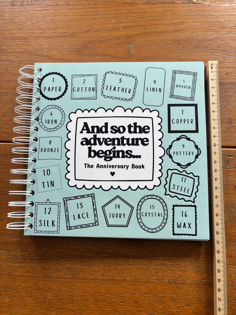 May include: A light blue spiral-bound anniversary book with the text "And so the adventure begins... The Anniversary Book" and sixteen numbered frames listing materials from 1 to 16. A wooden ruler shows the book is 28 cm in height.