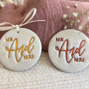 May include: Two white, circular wedding favours with the words "Mr And Mrs" in gold and copper script. Each favour has a small hole at the top with a white ribbon. The favours are on a pink cloth.