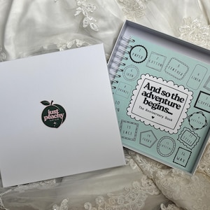 May include: A white square gift box with a peach logo and the text 'just peachy' next to an open spiral-bound anniversary book. The book cover is light blue with the text 'And so the adventure begins... The Anniversary Book' and numbered boxes.