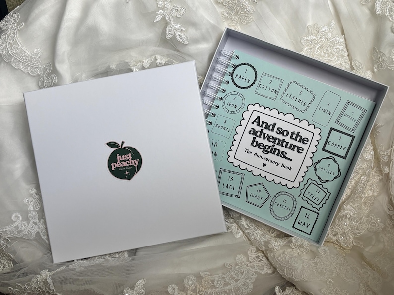 May include: A white square gift box with a peach logo and the text 'just peachy' next to an open spiral-bound anniversary book. The book cover is light blue with the text 'And so the adventure begins... The Anniversary Book' and numbered boxes.