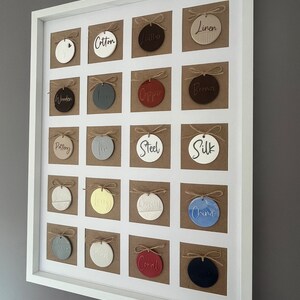 May include: A white frame with 20 round ceramic ornaments, each with a different color and word printed on it. The words include "Cotton", "Leather", "Linen", "Wooden", "Iron", "Copper", "Bronze", "Pottery", "Tin", "Steel", "Silk", "Silver", "Wool", "Crystal", "China", "Coral", "Black", "Gold", "White", and "Grey".