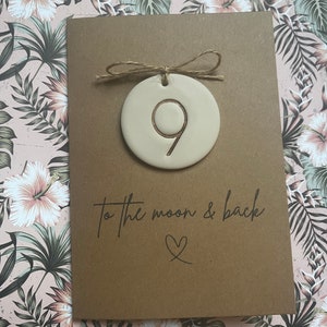 May include: A brown card with the text "to the moon & back" and a heart symbol. A small, round, beige ceramic ornament with the number 9 is attached to the card with twine.